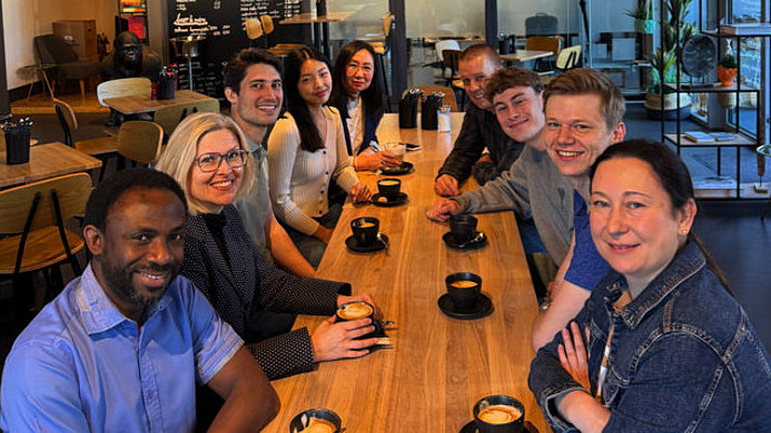 Gruppe von Menschen sitzt an einem langen Holztisch in einem Café mit runden, bunten Deckenlampen und trinkt Kaffee