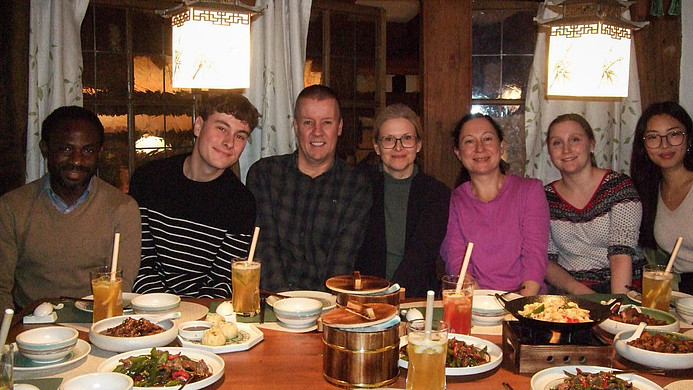 Gruppe von sechs Personen sitzt an einem Tisch mit asiatisch anmutenden Speisen und Getränken in einem Restaurant mit hölzernen Wänden und dekorativen Lampen