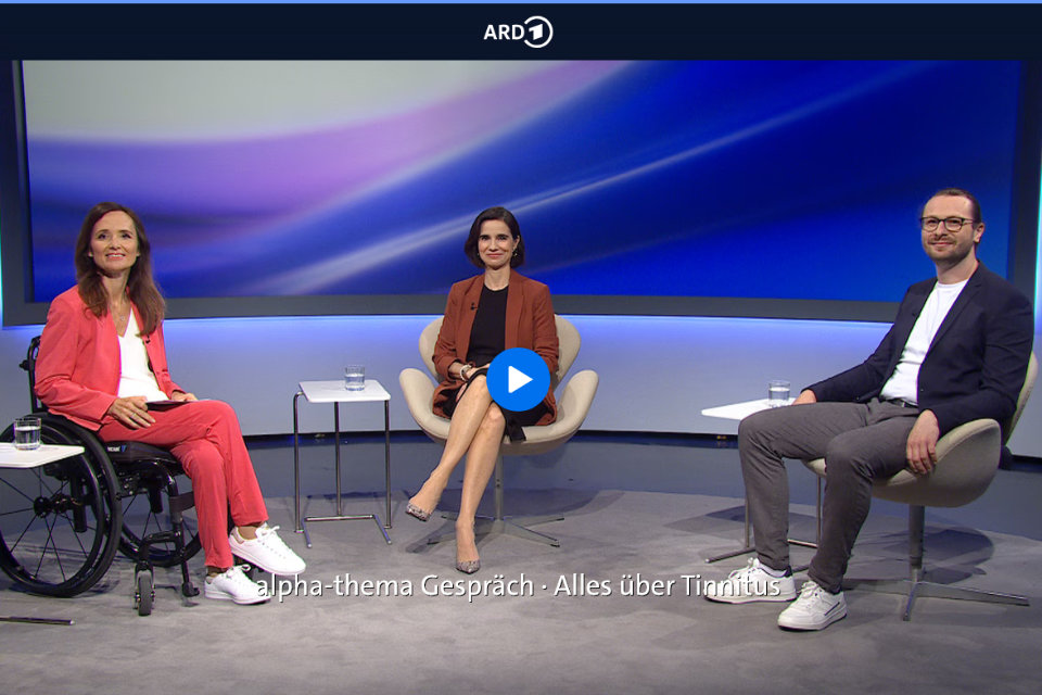 Drei Personen sitzen in einem Fernsehstudio auf Stühlen, eine Frau links im Rollstuhl trägt einen roten Anzug, die mittlere Frau trägt ein braunes Jackett und Rock, der Mann rechts trägt ein dunkles Jackett und helle Hose